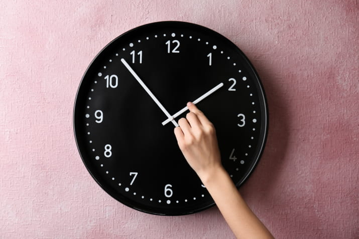 A hand changing the time on a black clock, which sits on a pink background