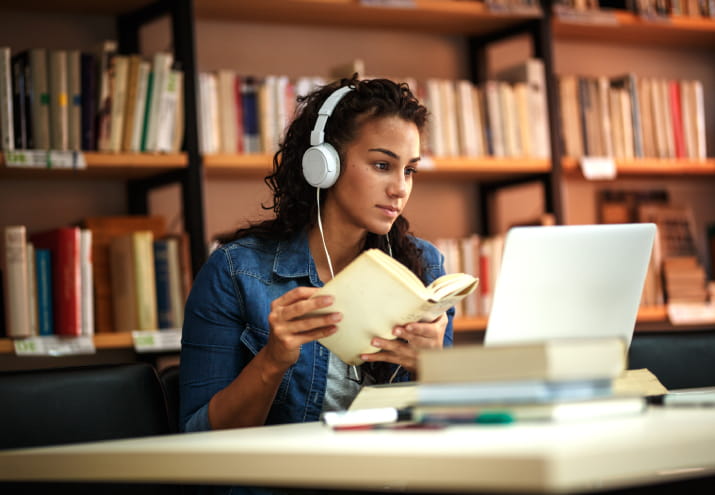 A young woman wearing headphones and writing notes while looking at a laptop
