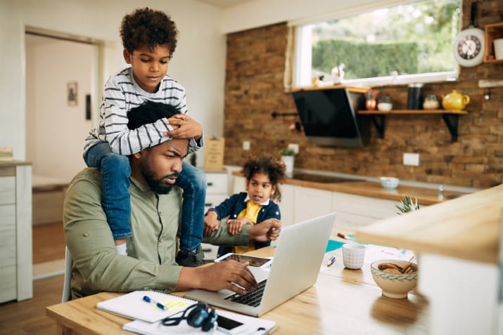 A man trying to work on a laptop with his son on his head and his daughter pulling his arm