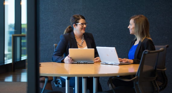 Two people in a workplace meeting, using laptop computers, seated at a table.